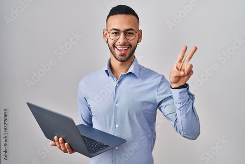 Young hispanic man working using computer laptop smiling with happy face winking at the camera doing victory sign with fingers. number two.