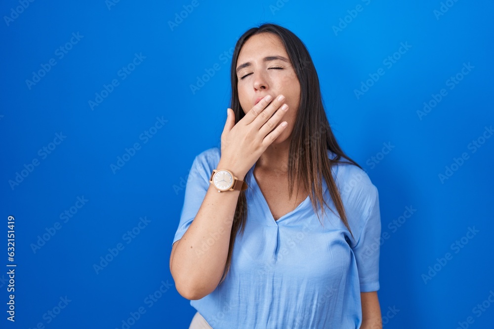 Young brunette woman standing over blue background bored yawning tired covering mouth with hand. restless and sleepiness. Young brunette woman standing over blue background bored yawning tired covering mouth with hand. restless and sleepiness.