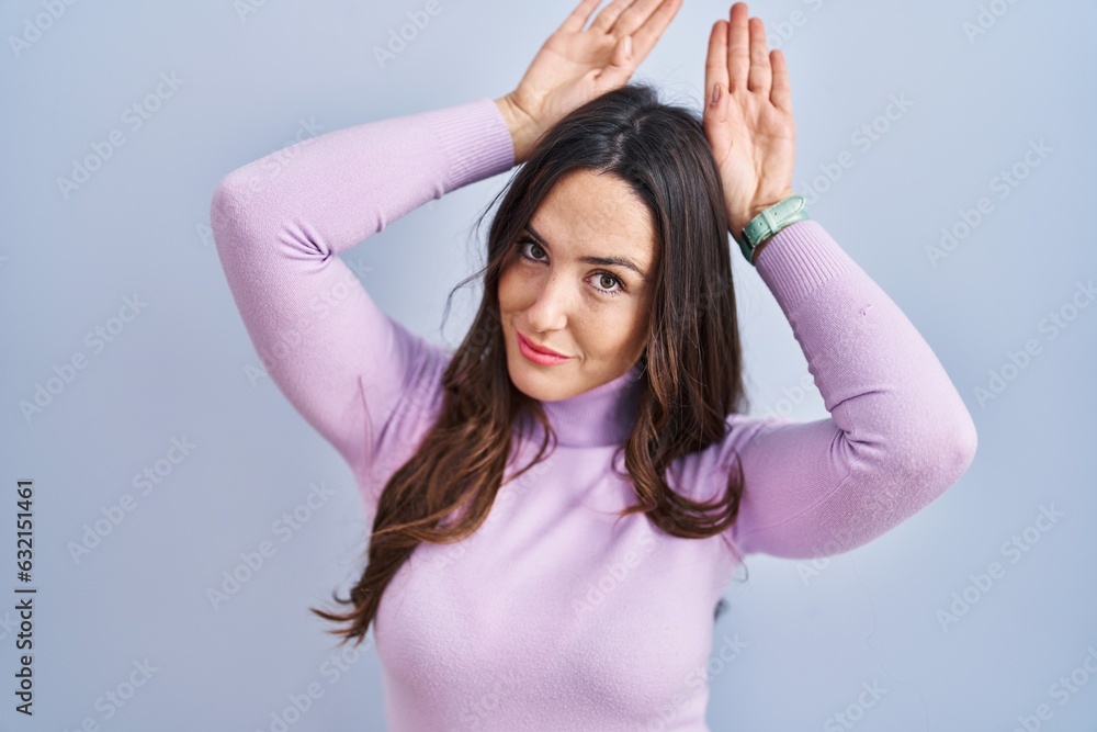 Young brunette woman standing over blue background doing bunny ears gesture with hands palms looking cynical and skeptical. easter rabbit concept.
