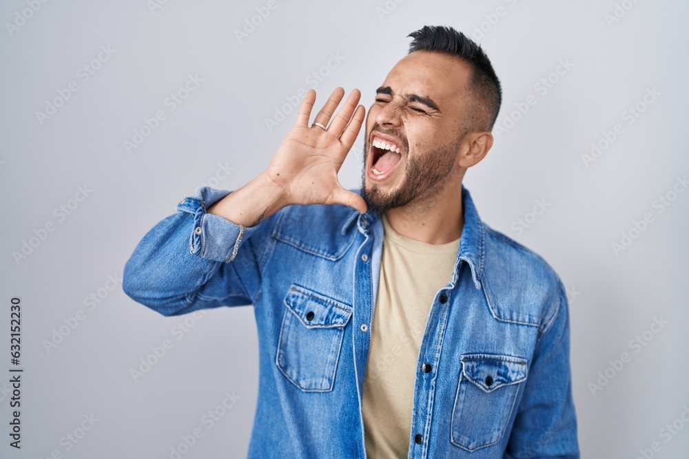 Young hispanic man standing over isolated background shouting and screaming loud to side with hand on mouth. communication concept. Young hispanic man standing over isolated background shouting and screaming loud to side with hand on mouth. communication concept.