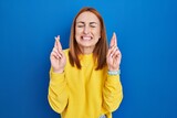 Young woman standing over blue background gesturing finger crossed smiling with hope and eyes closed. luck and superstitious concept.