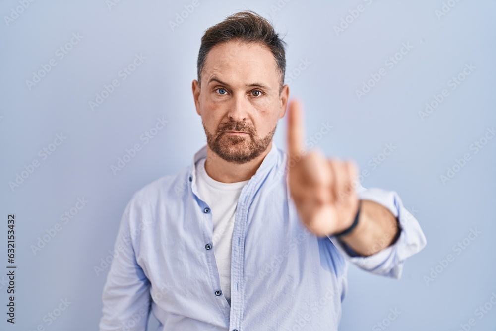 Middle age caucasian man standing over blue background pointing with finger up and angry expression, showing no gesture Middle age caucasian man standing over blue background pointing with finger up and angry expression, showing no gesture