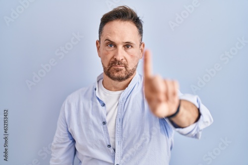 Middle age caucasian man standing over blue background pointing with finger up and angry expression, showing no gesture