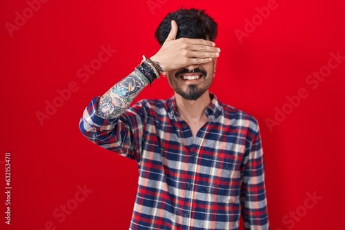 Young hispanic man with beard standing over red background smiling and laughing with hand on face covering eyes for surprise. blind concept.