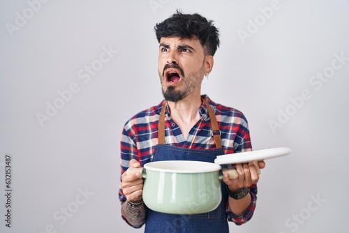 Slika na platnu Young hispanic man with beard wearing apron holding cooking pot angry and mad screaming frustrated and furious, shouting with anger