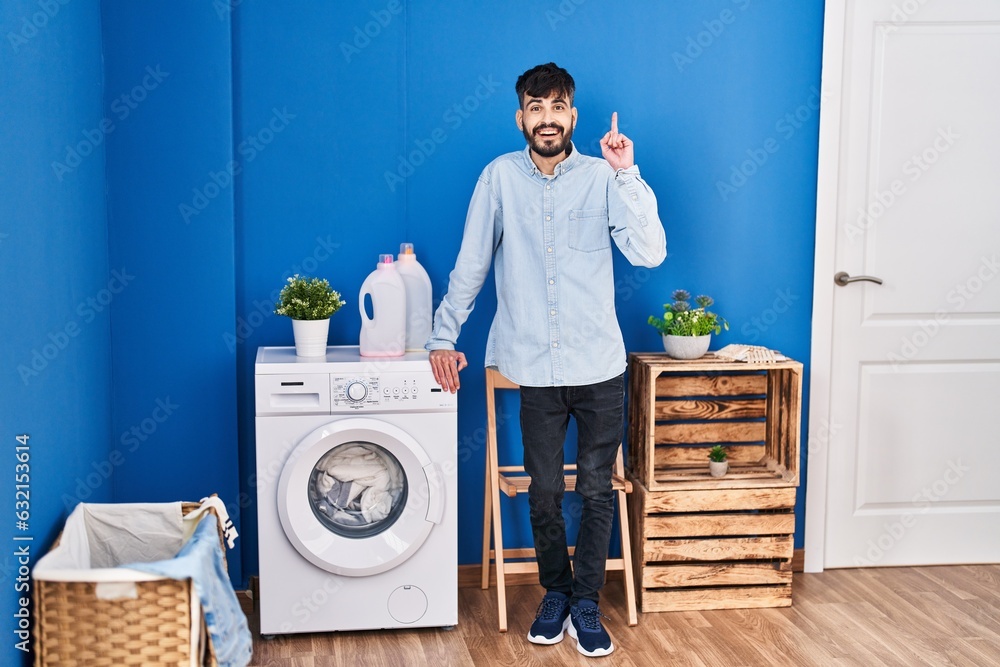 Young hispanic man with beard doing laundry standing at laundry room surprised with an idea or question pointing finger with happy face, number one Young hispanic man with beard doing laundry standing at laundry room surprised with an idea or question pointing finger with happy face, number one