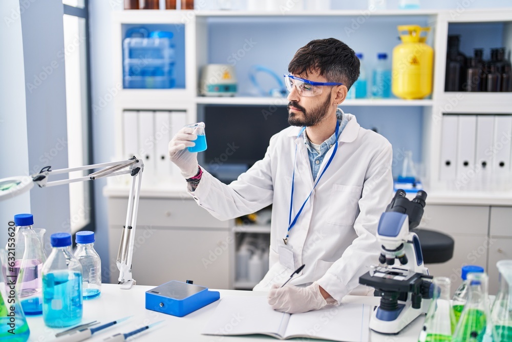 Young hispanic man scientist writing on notebook measuring liquid at laboratory Young hispanic man scientist writing on notebook measuring liquid at laboratory