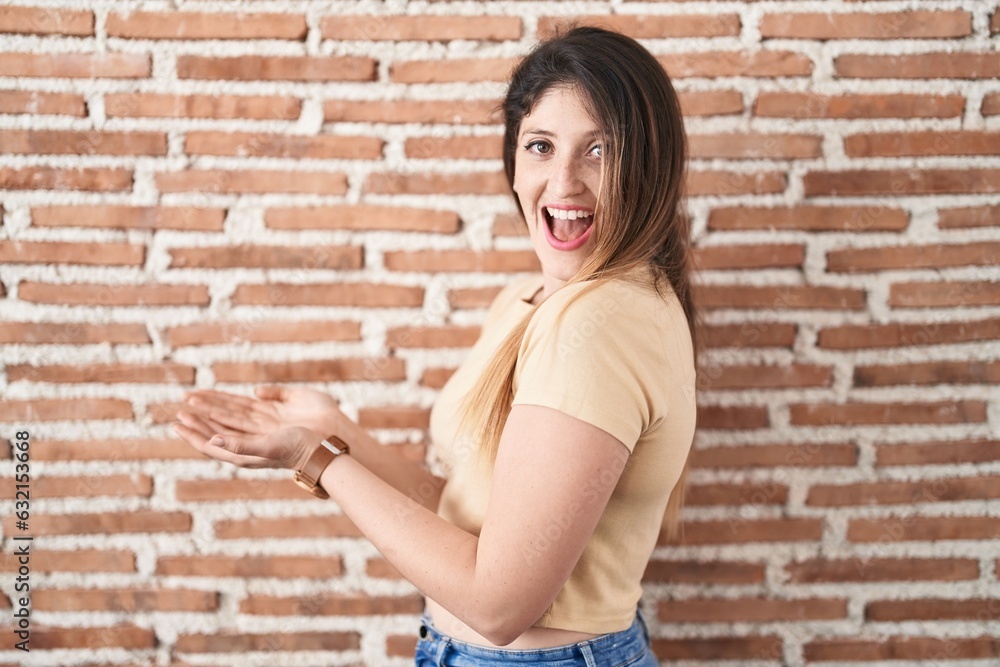 Young brunette woman standing over bricks wall pointing aside with hands open palms showing copy space, presenting advertisement smiling excited happy Young brunette woman standing over bricks wall pointing aside with hands open palms showing copy space, presenting advertisement smiling excited happy