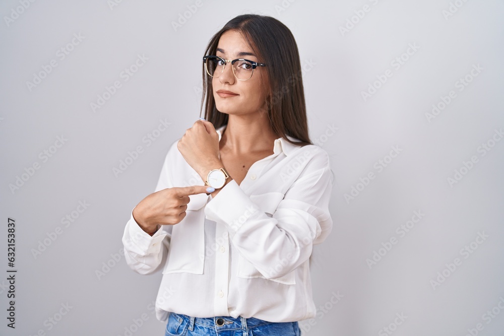 Young brunette woman wearing glasses in hurry pointing to watch time, impatience, looking at the camera with relaxed expression Young brunette woman wearing glasses in hurry pointing to watch time, impatience, looking at the camera with relaxed expression