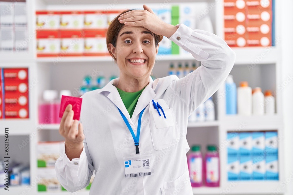 Brunette woman working at pharmacy drugstore holding condom stressed and frustrated with hand on head, surprised and angry face