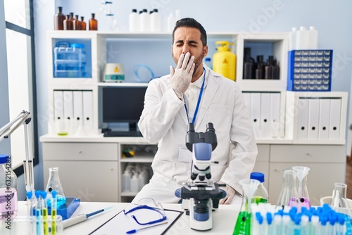 Young hispanic man with beard working at scientist laboratory bored yawning tired covering mouth with hand. restless and sleepiness.