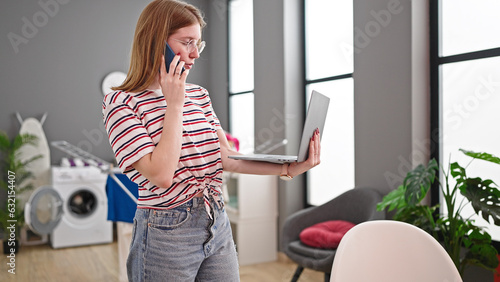 Young blonde woman using laptop talking on smartphone at dinning room