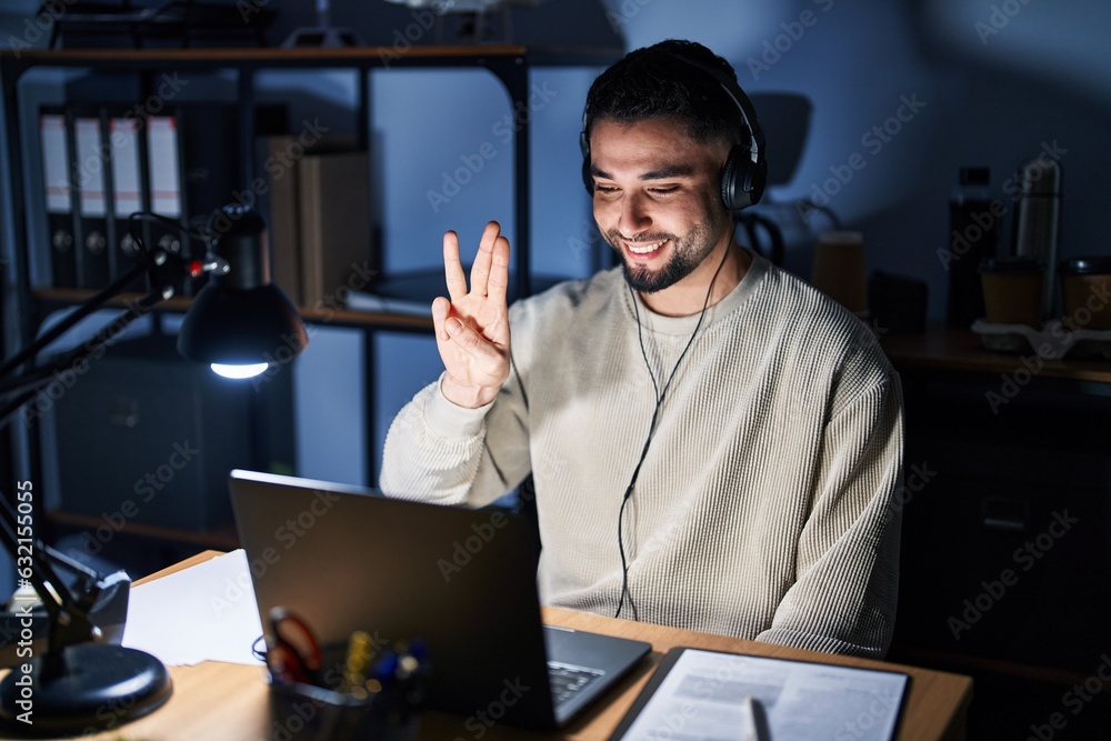 Young handsome man working using computer laptop at night showing and pointing up with fingers number three while smiling confident and happy. Young handsome man working using computer laptop at night showing and pointing up with fingers number three while smiling confident and happy.