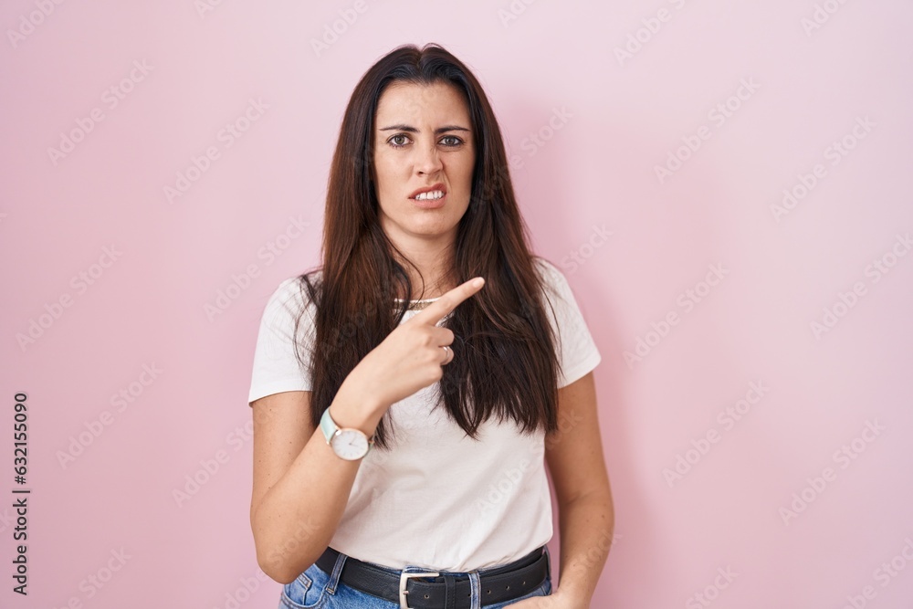 Young brunette woman standing over pink background pointing aside worried and nervous with forefinger, concerned and surprised expression Young brunette woman standing over pink background pointing aside worried and nervous with forefinger, concerned and surprised expression