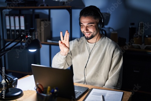 Young handsome man working using computer laptop at night showing and pointing up with fingers number three while smiling confident and happy.