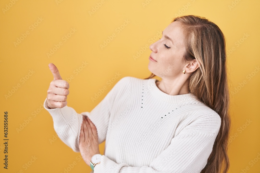 Young caucasian woman wearing white sweater over yellow background looking proud, smiling doing thumbs up gesture to the side Young caucasian woman wearing white sweater over yellow background looking proud, smiling doing thumbs up gesture to the side