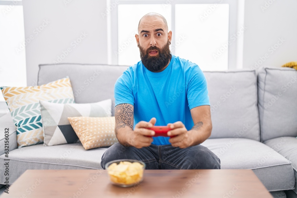 Young hispanic man with beard and tattoos playing video game sitting on the sofa in shock face, looking skeptical and sarcastic, surprised with open mouth Young hispanic man with beard and tattoos playing video game sitting on the sofa in shock face, looking skeptical and sarcastic, surprised with open mouth