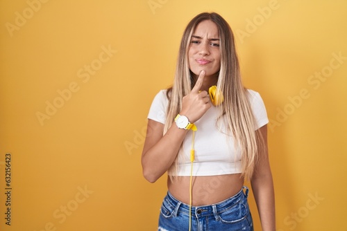 Young blonde woman standing over yellow background wearing headphones thinking concentrated about doubt with finger on chin and looking up wondering