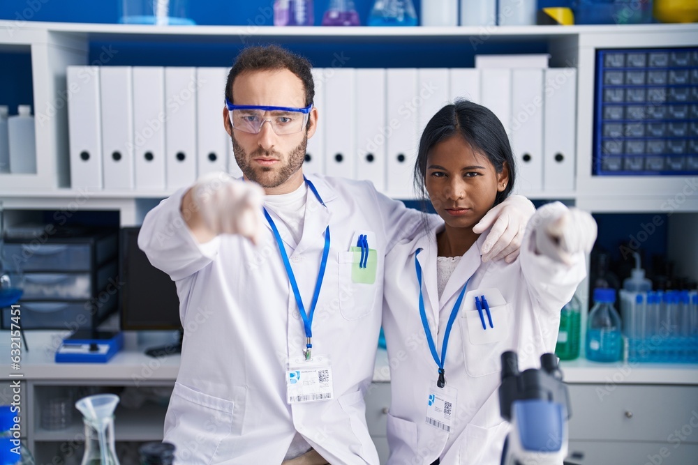 Interracial couple working at scientist laboratory pointing with finger to the camera and to you, confident gesture looking serious Interracial couple working at scientist laboratory pointing with finger to the camera and to you, confident gesture looking serious