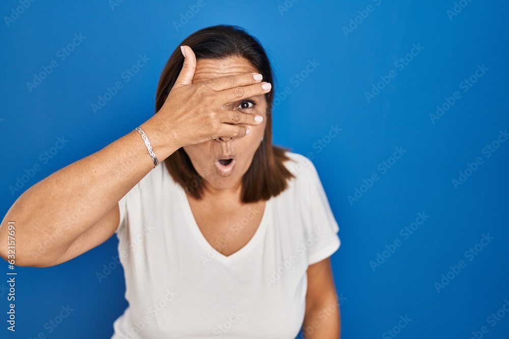 Hispanic mature woman standing over blue background peeking in shock covering face and eyes with hand, looking through fingers with embarrassed expression. Hispanic mature woman standing over blue background peeking in shock covering face and eyes with hand, looking through fingers with embarrassed expression.