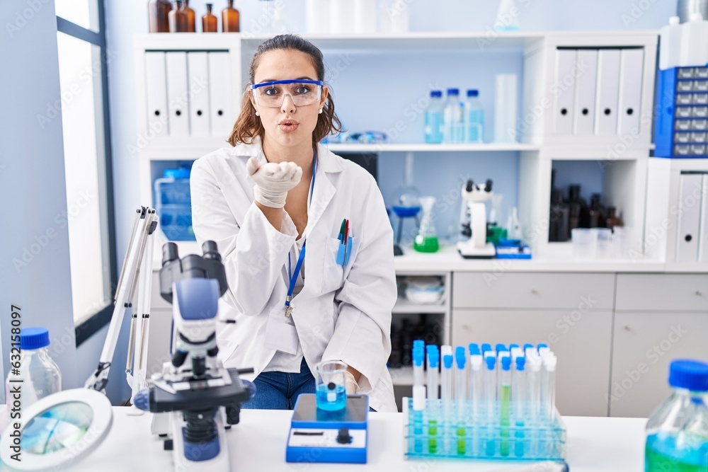 Young hispanic woman working at scientist laboratory looking at the camera blowing a kiss with hand on air being lovely and sexy. love expression. Young hispanic woman working at scientist laboratory looking at the camera blowing a kiss with hand on air being lovely and sexy. love expression.