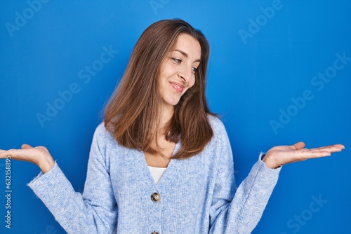 Young woman standing over blue background smiling showing both hands open palms, presenting and advertising comparison and balance