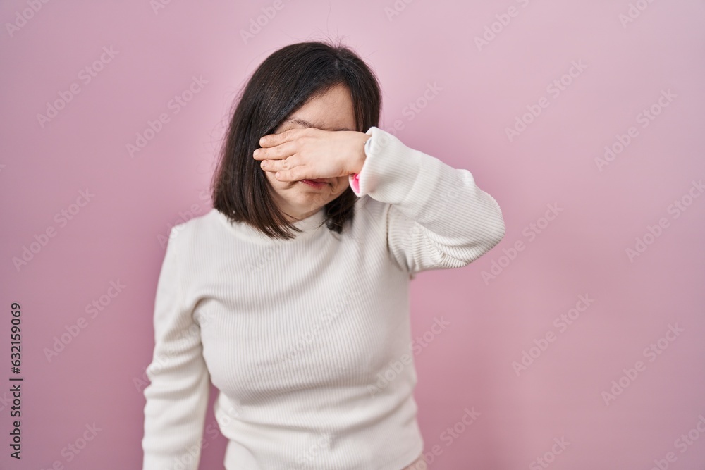 Woman with down syndrome standing over pink background covering eyes with hand, looking serious and sad. sightless, hiding and rejection concept Woman with down syndrome standing over pink background covering eyes with hand, looking serious and sad. sightless, hiding and rejection concept