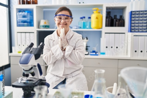 Hispanic girl with down syndrome working at scientist laboratory praying with hands together asking for forgiveness smiling confident.
