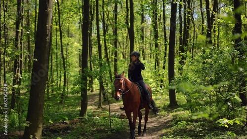 Wide shot green spring summer forest in sunshine with graceful stallion walking with equestrian on back and leaving. Confident purebred horse and Caucasian pretty woman passing in sunshine