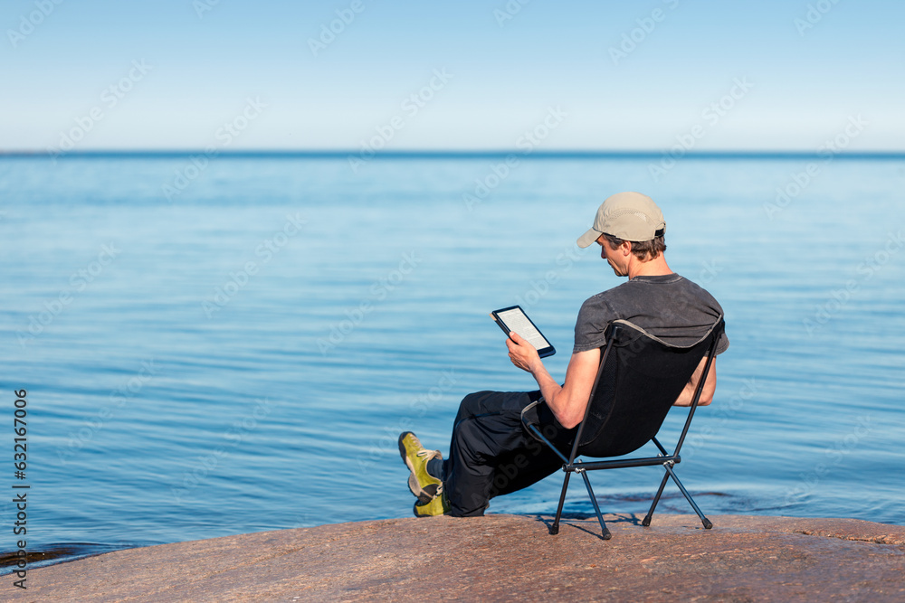 A man is reading an e-book on the seashore sitting in a camping chair.