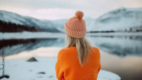 Young blond woman in orange sweater and winter hat staring at the lake