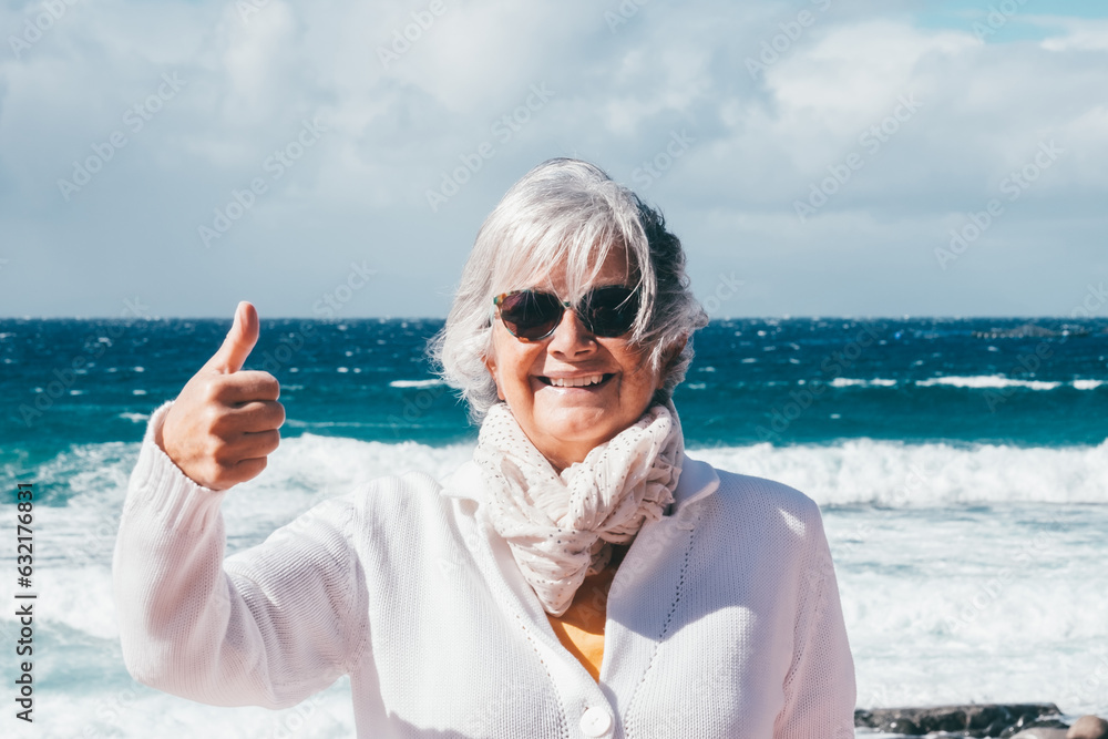Happy senior white-haired woman in outdoors enjoying sea vacation, elderly smiling lady in a windy day at the beach looking at camera with thumb up
