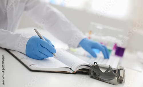 Employee of the chemical laboratory teacher chemist holds silvery pen in his hand makes notes in diary records. Test data from reactions examining test tubes with the substance arm in gloves