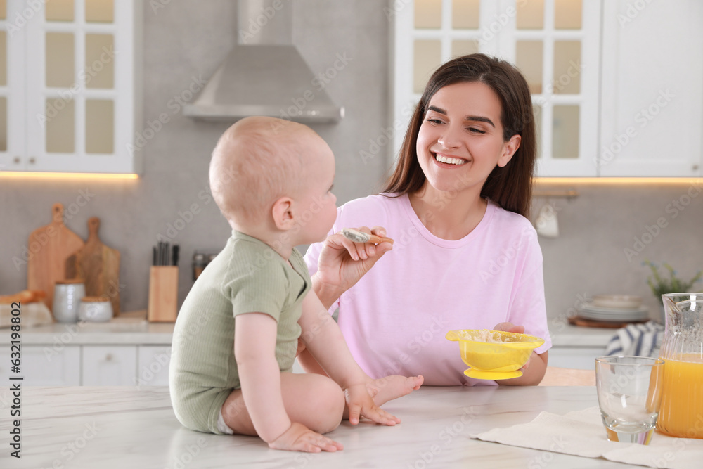 Happy young woman feeding her cute little baby at table in kitchen Happy young woman feeding her cute little baby at table in kitchen