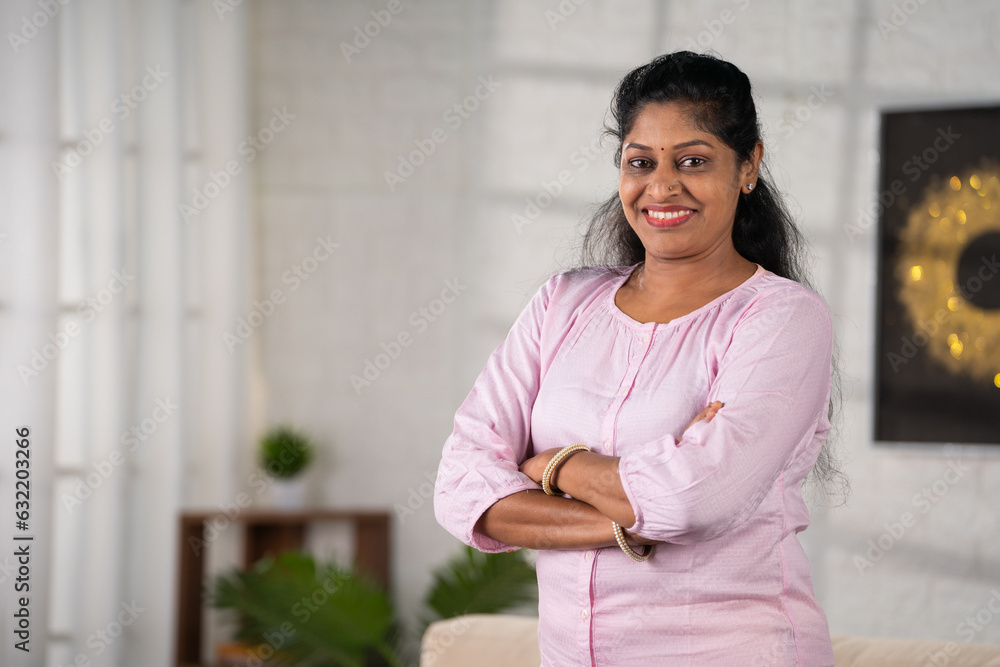 Happy smiling middle aged indian woman standing with arms crossed by looking camera at home with copy space - concept of successful, empowerment and inspirational. Happy smiling middle aged indian woman standing with arms crossed by looking camera at home with copy space - concept of successful, empowerment and inspirational.