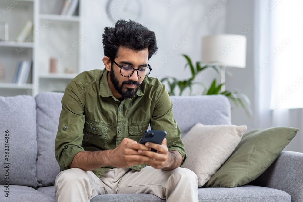 Serious thinking man sitting on sofa in living room at home, Indian man reading news on phone, using app on smartphone, browsing internet pages. Serious thinking man sitting on sofa in living room at home, Indian man reading news on phone, using app on smartphone, browsing internet pages.