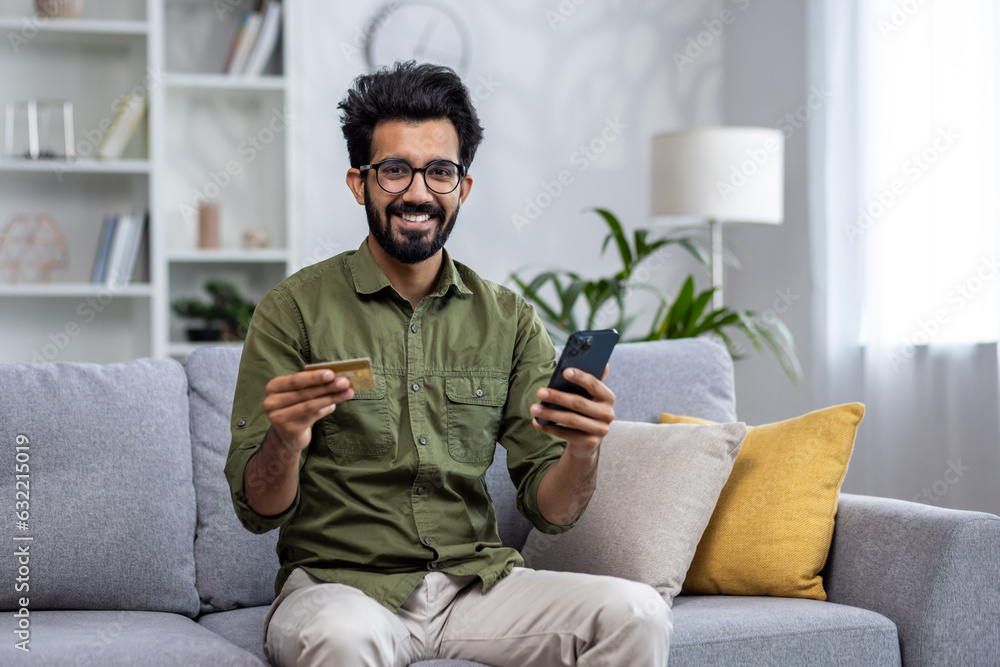 Portrait of happy satisfied online shopper, hispanic woman smiling and looking at camera sitting on sofa in living room at home, man holding bank credit card and phone. Portrait of happy satisfied online shopper, hispanic woman smiling and looking at camera sitting on sofa in living room at home, man holding bank credit card and phone.