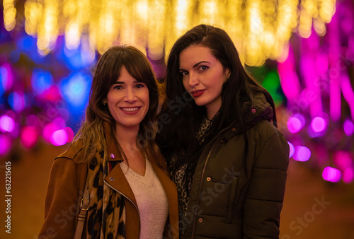 couple of beautiful girls dressed in coats posing at night in a place full of coloured Christmas lights in the background out of focus.