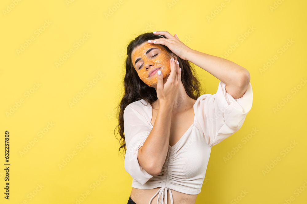A young brunette girl with a cleansing scrub mask on her face, looks at the camera and smiles, against a yellow background. A young brunette girl with a cleansing scrub mask on her face, looks at the camera and smiles, against a yellow background.