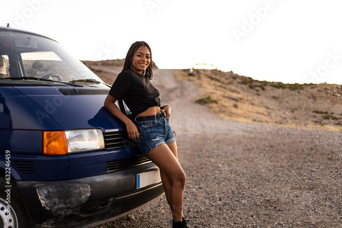 A Latina girl with braided hair in casual clothes is leaning on a camper van while looking at the camera happily. Concept of African women traveling in summer to the mountains. Copyspace.