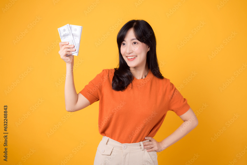 Vivacious young Asian woman in her 30s, holding cash money dollars, standing on vibrant yellow backdrop. Vivacious young Asian woman in her 30s, holding cash money dollars, standing on vibrant yellow backdrop.