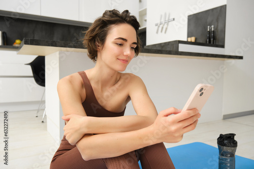 Portrait of sporty girl with mobile phone, sitting on yoga mat, using smartphone during workout at home, watching fitness turorial on telephone photo