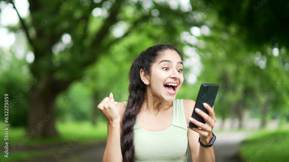 Happy excited young sports woman checking results of training while looking at smartphone in urban city park. Smiling female happy with good successful workout results or great news on mobile phone Happy excited young sports woman checking results of training while looking at smartphone in urban city park. Smiling female happy with good successful workout results or great news on mobile phone