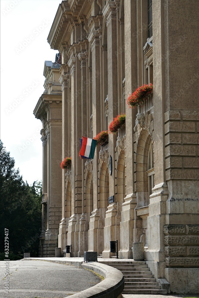 Węgierska flaga powiewa na Uniwersytecie w Debreczynie. Na budynku widać liczne zdobienia i czerwone kwiaty. - obrazy, fototapety, plakaty 
