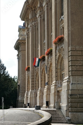 Węgierska flaga powiewa na Uniwersytecie w Debreczynie. Na budynku widać liczne zdobienia i czerwone kwiaty.