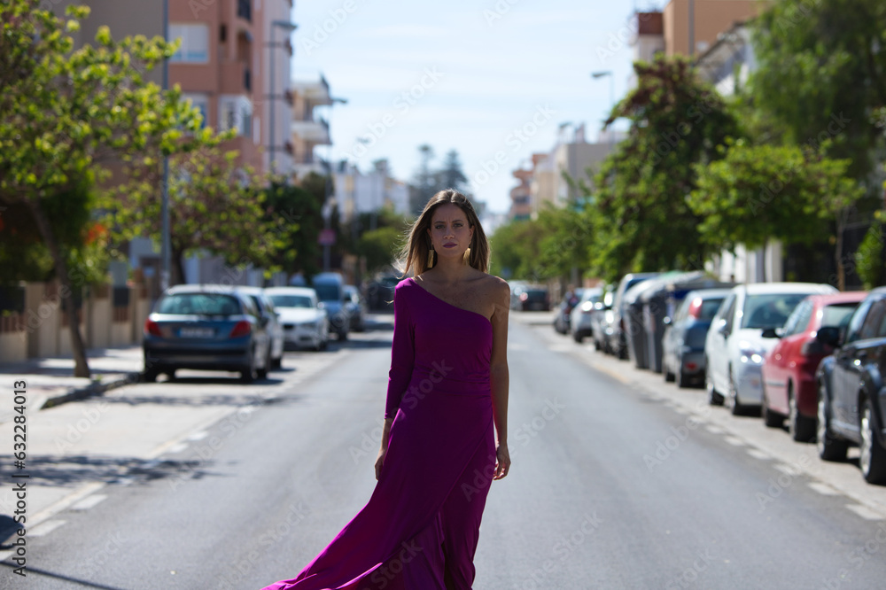 Fototapeta premium Young beautiful blonde woman dressed in purple dress walks along a large lonely avenue among the stopped cars. The woman makes different body expressions while parading like a model.