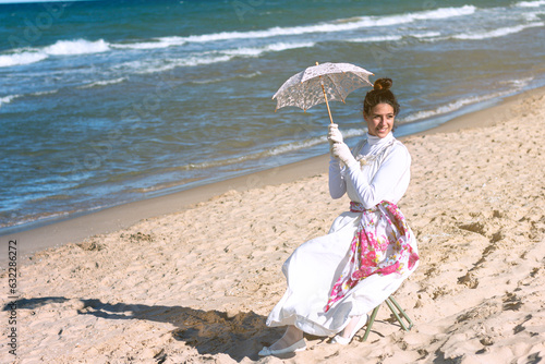 girl sitting on the shore of the beach in period costume with umbrella and hat photo