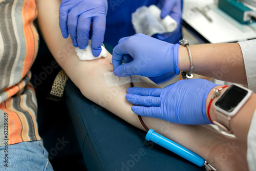 blood donation, two nurses in blue gloves connect the collection system with a needle to the patient's left arm, the donor is sitting in a chair in a medical facility, no face, medium close-up