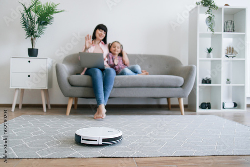 Selective focus of modern robotic vacuum keeping floor tidy while female owners starting video call on gadget. Mother-daughter duo enjoying online communication while wireless appliance doing job.