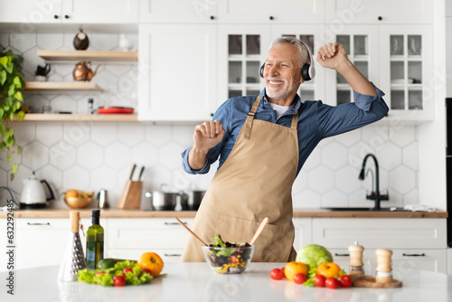 Positive Senior Man In Headphones Listening Music While Cooking Food In Kitchen